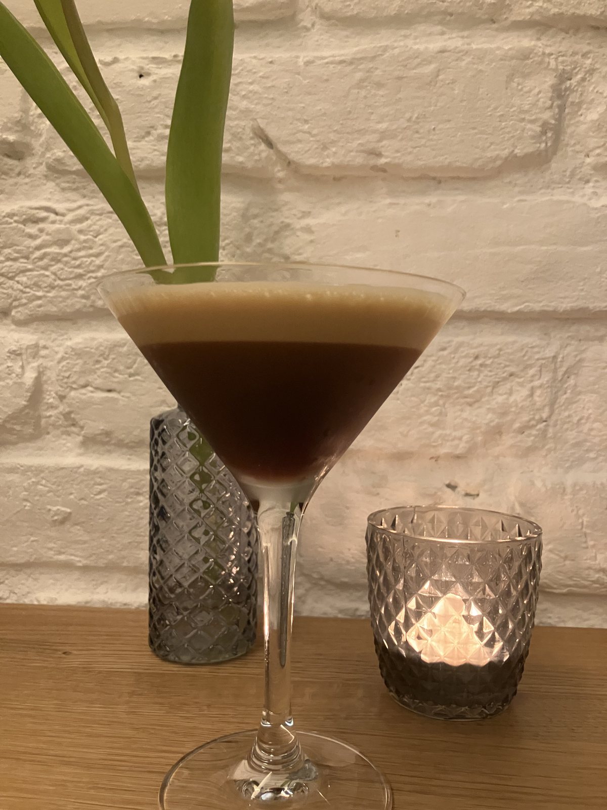 Espresso Martini im Stielglas vor der weiß gekalkten Ziegelwand, daneben ein Windlicht